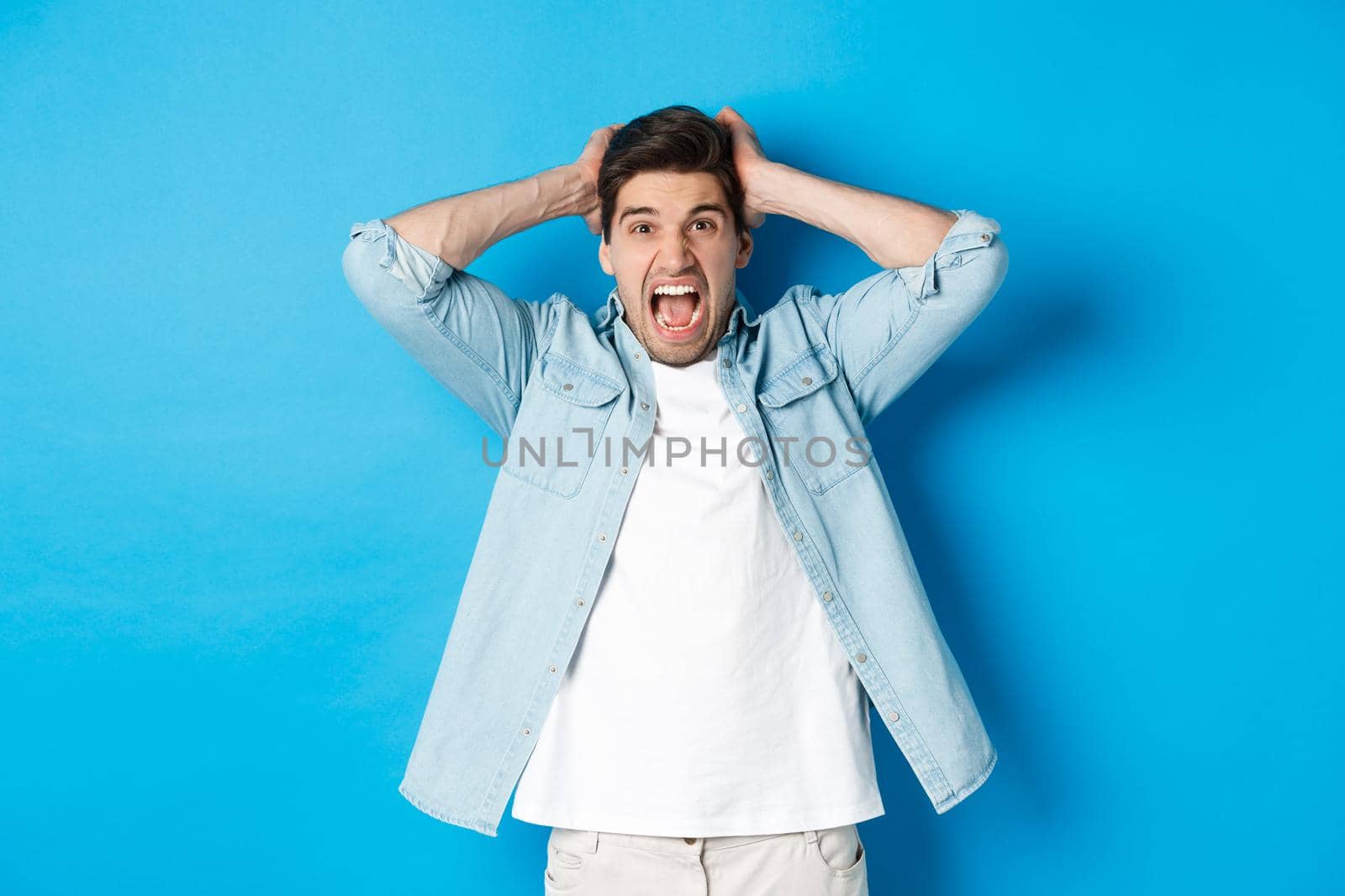 Frustrated guy losing, yelling displeased and looking at camera, holding hands on head, standing over blue background.