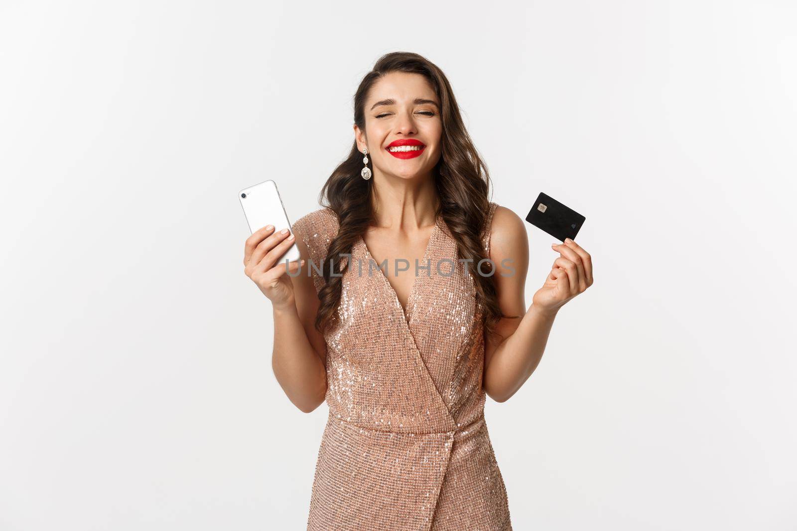 Online shopping and holidays concept. Satisfied and happy woman in elegant dress smiling, using credit card and mobile phone, standing over white background by Benzoix
