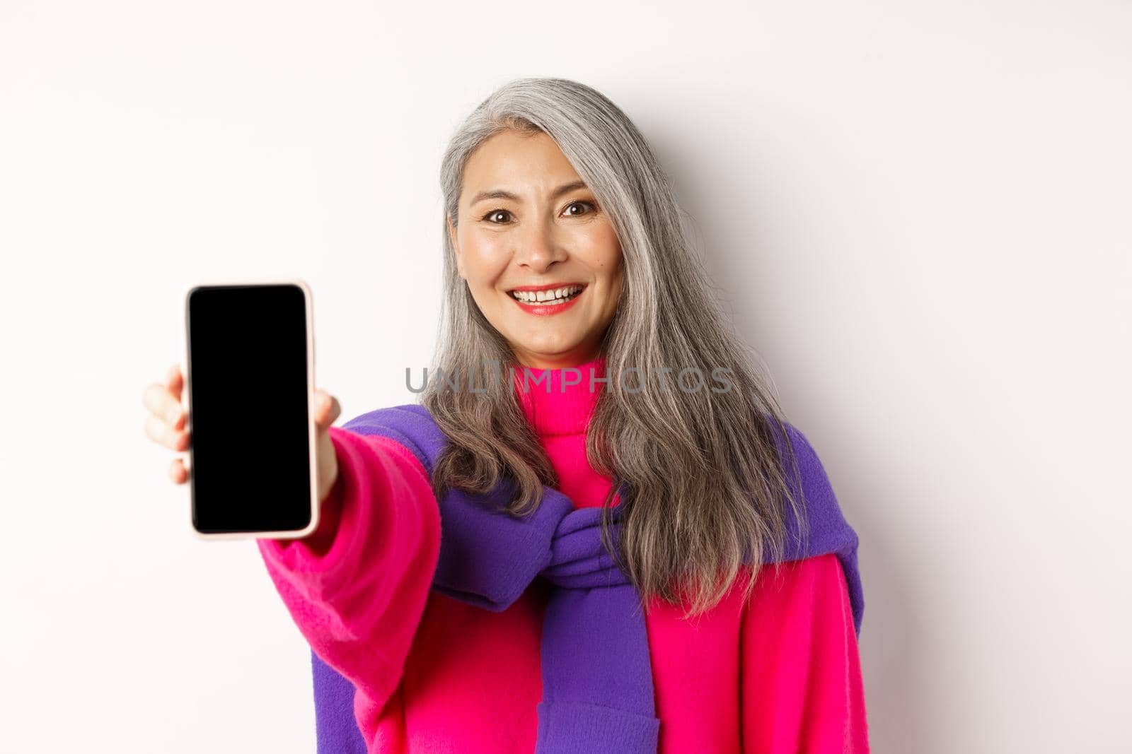 Online shopping. Close up of stylish asian senior woman extending hand with mobile phone, showing blank smartphone screen and smiling, standing over white background by Benzoix