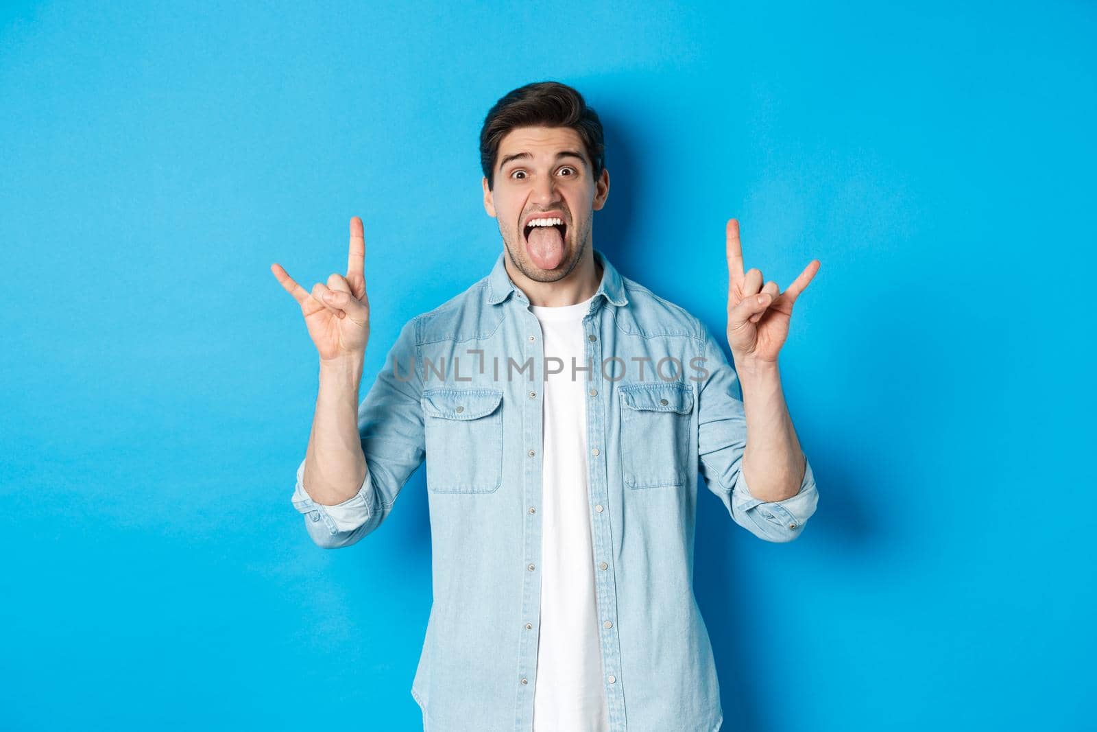 Cool guy having fun, enjoying concert, showing rock sign and tongue, standing over blue background by Benzoix