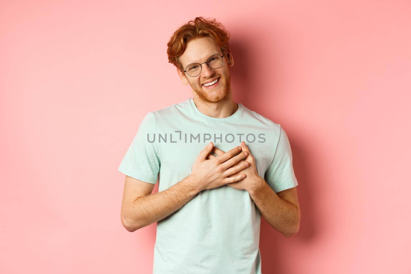 Attractive young man with ginger hair, holding hands on heart and smiling grateful, saying thank you, express gratitude, standing over pink background by Benzoix