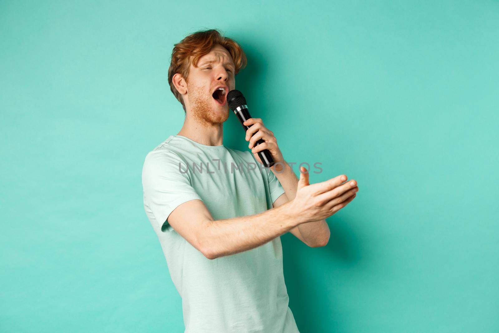 Passionate redhead man in t-shirt singing serenade with microphone, looking aside at karaoke and gesturing, standing over mint background by Benzoix