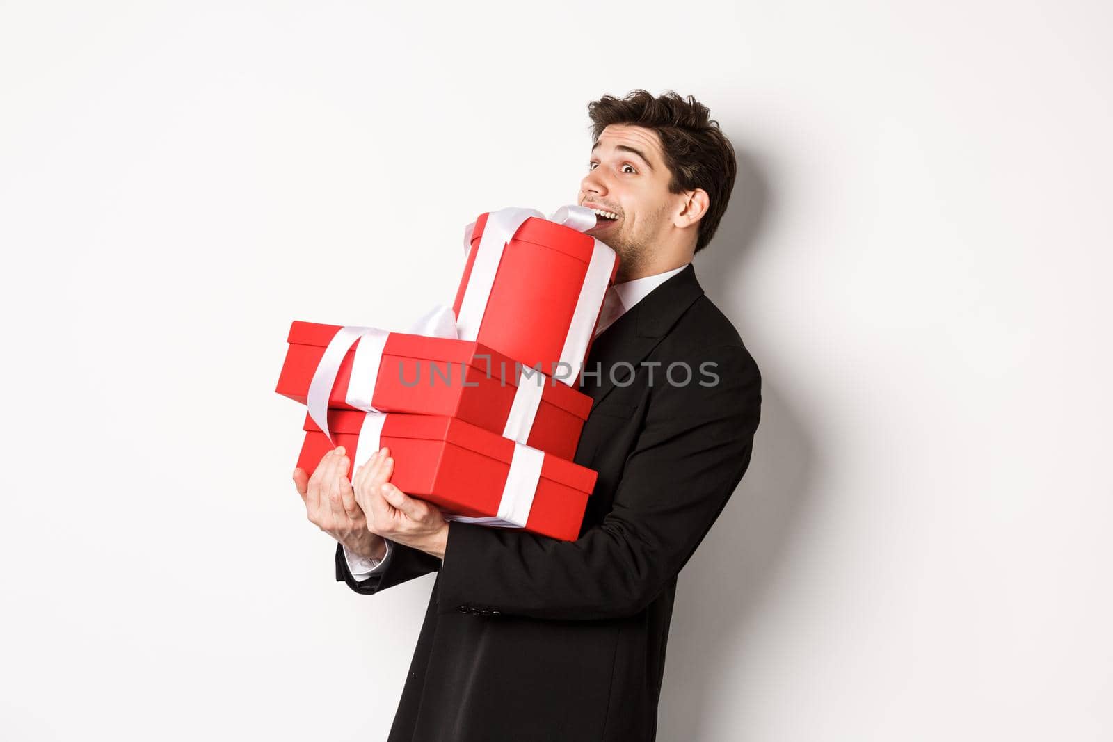 Concept of christmas holidays, celebration and lifestyle. Image of excited handsome man buying gifts for new year, standing happy over white background in black suit by Benzoix