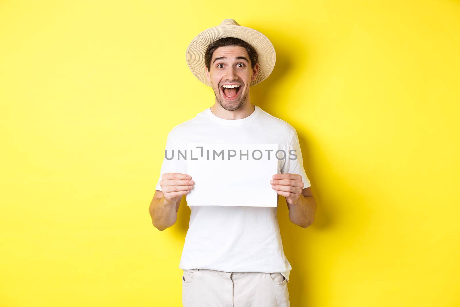 Excited tourist showing your logo or sign on blank piece of paper, smiling amazed, standing against yellow background by Benzoix