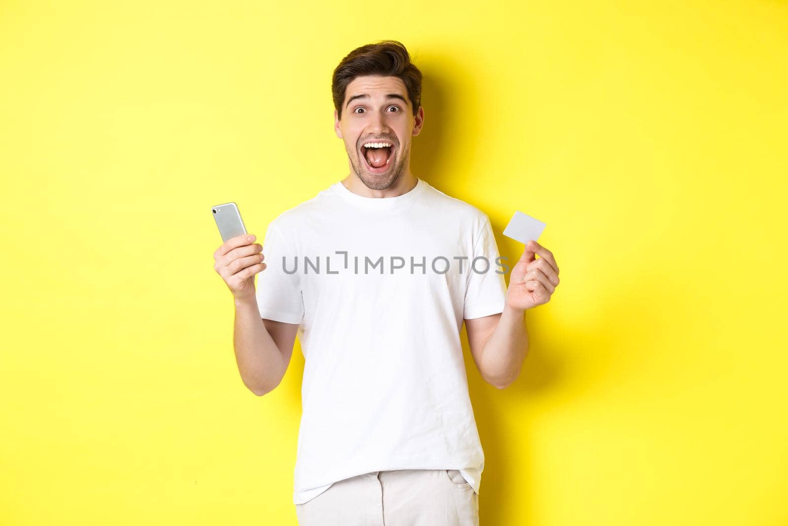 Surprised guy holding smartphone and credit card, online shopping on black friday, standing over yellow background.