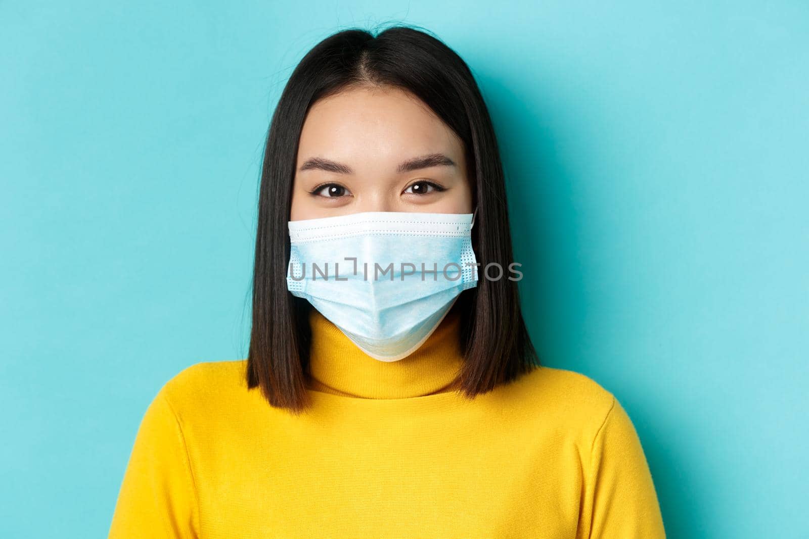 Covid-19, social distancing and pandemic concept. Close up of young asian woman with short dark hair, wearing medical mask and smiling with eyes, looking hopeful at camera by Benzoix