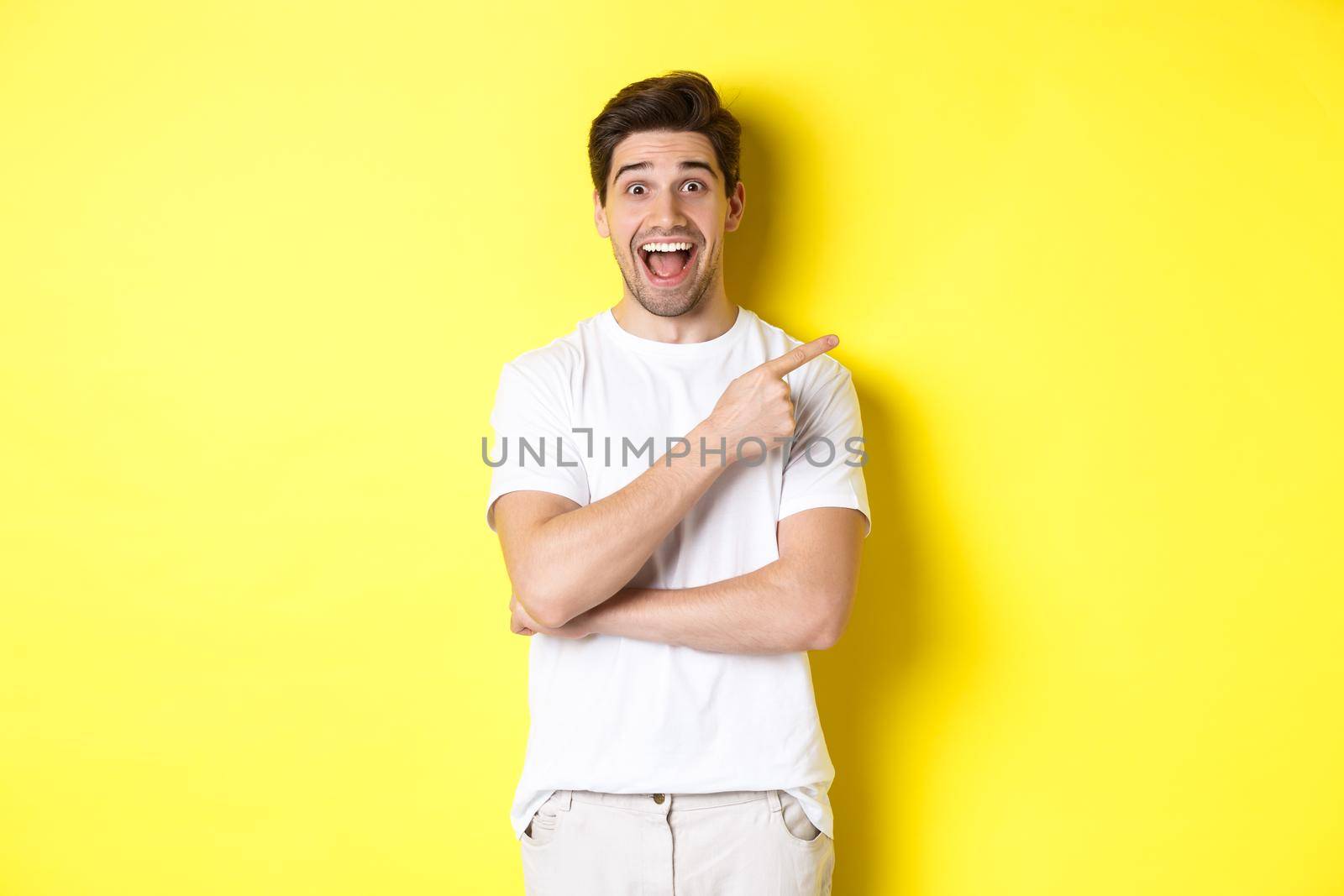 Image of excited smiling man showing black friday offers, pointing finger right and looking amazed, standing over yellow background.