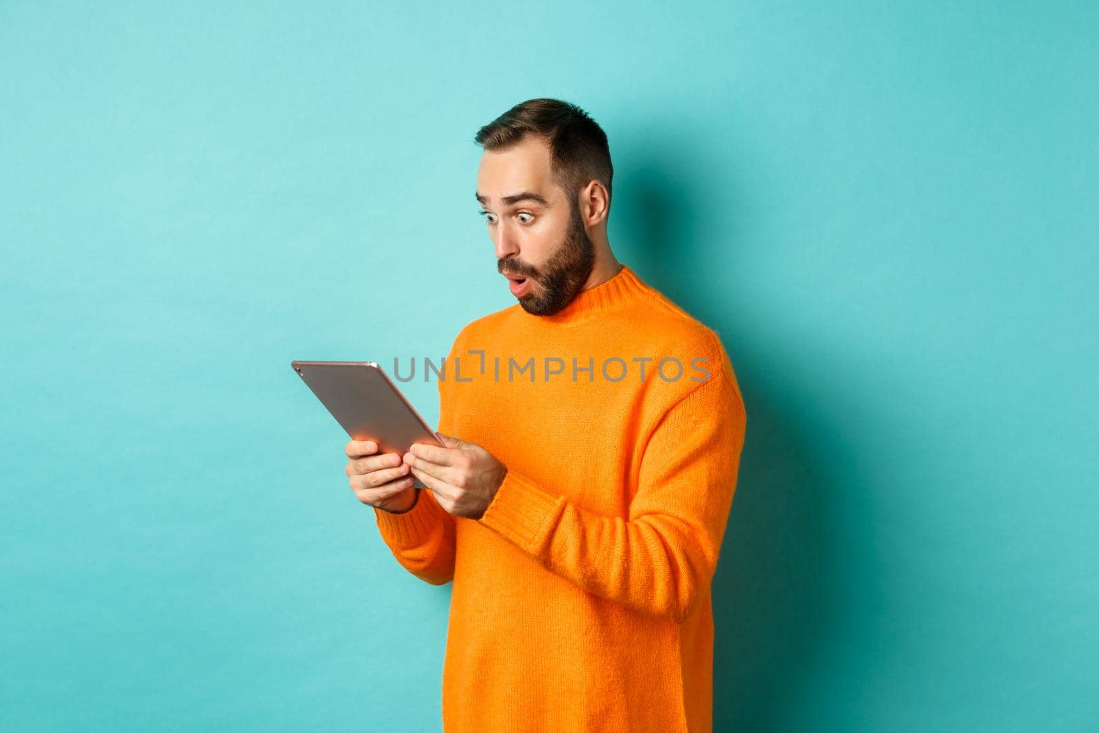 Image of male model in orange sweater staring at digital tablet screen, looking surprised, standing over light blue background.