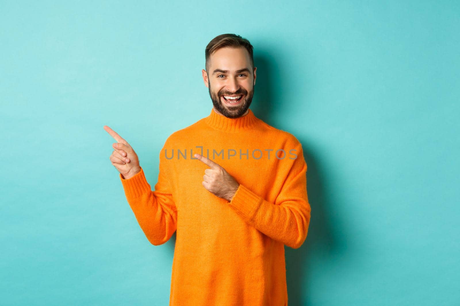 Handsome smiling man pointing fingers left, showing your logo, standing in winter orange sweater, turquoise background.