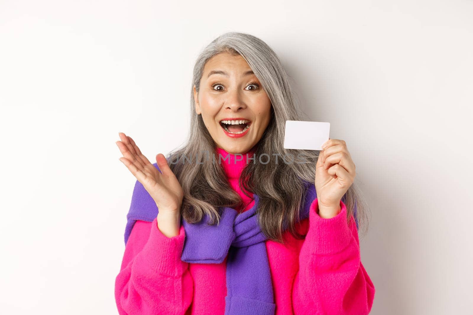 Shopping concept. Happy asian old aldy looking impressed and showing plastic credit card of her bank, standing over white background by Benzoix