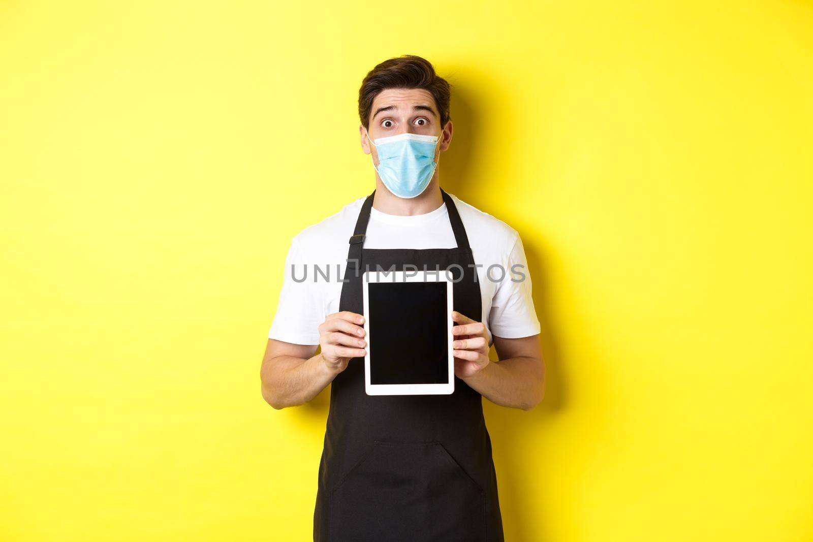 Concept of covid-19, small business and pandemic. Surprised waiter in black apron and medical mask showing tablet screen, standing over yellow background by Benzoix