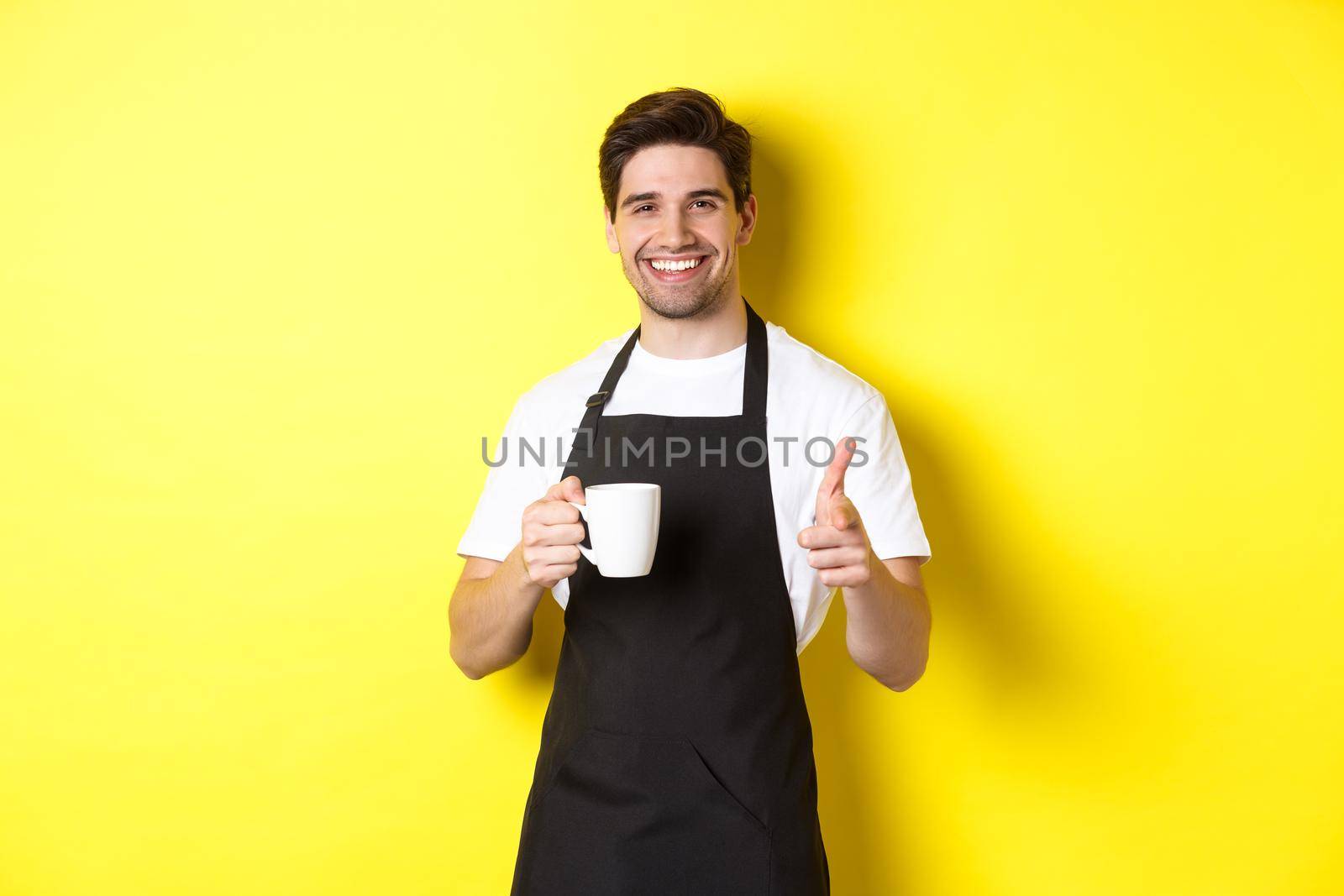 Barista bringing coffee and pointing finger gun at camera, standing in black apron against yellow background.