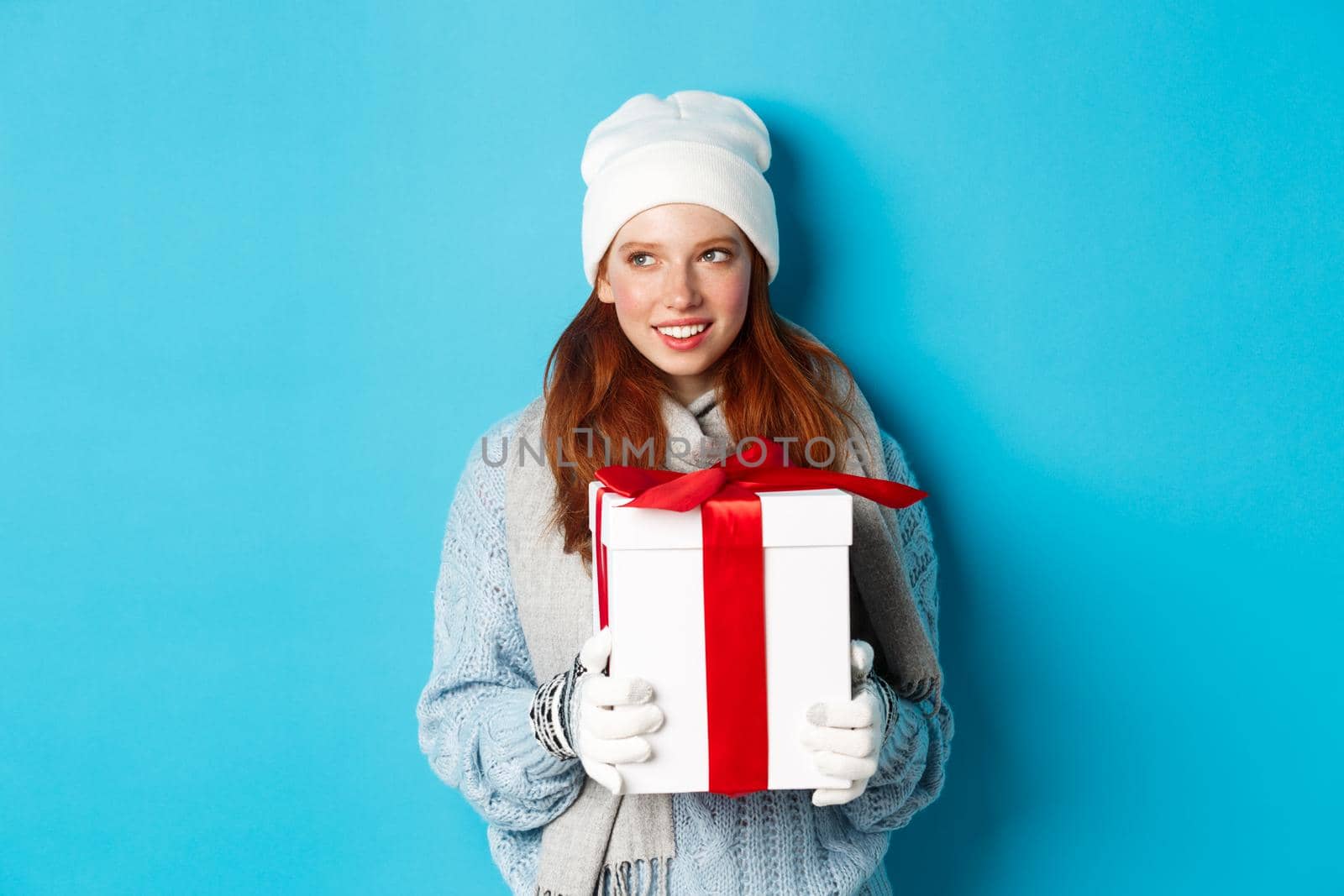 Winter holidays and Christmas sales concept. Thoughtful redhead woman standing in white beanie and gloves, holding holiday gift and looking left, thinking, standing over blue background.