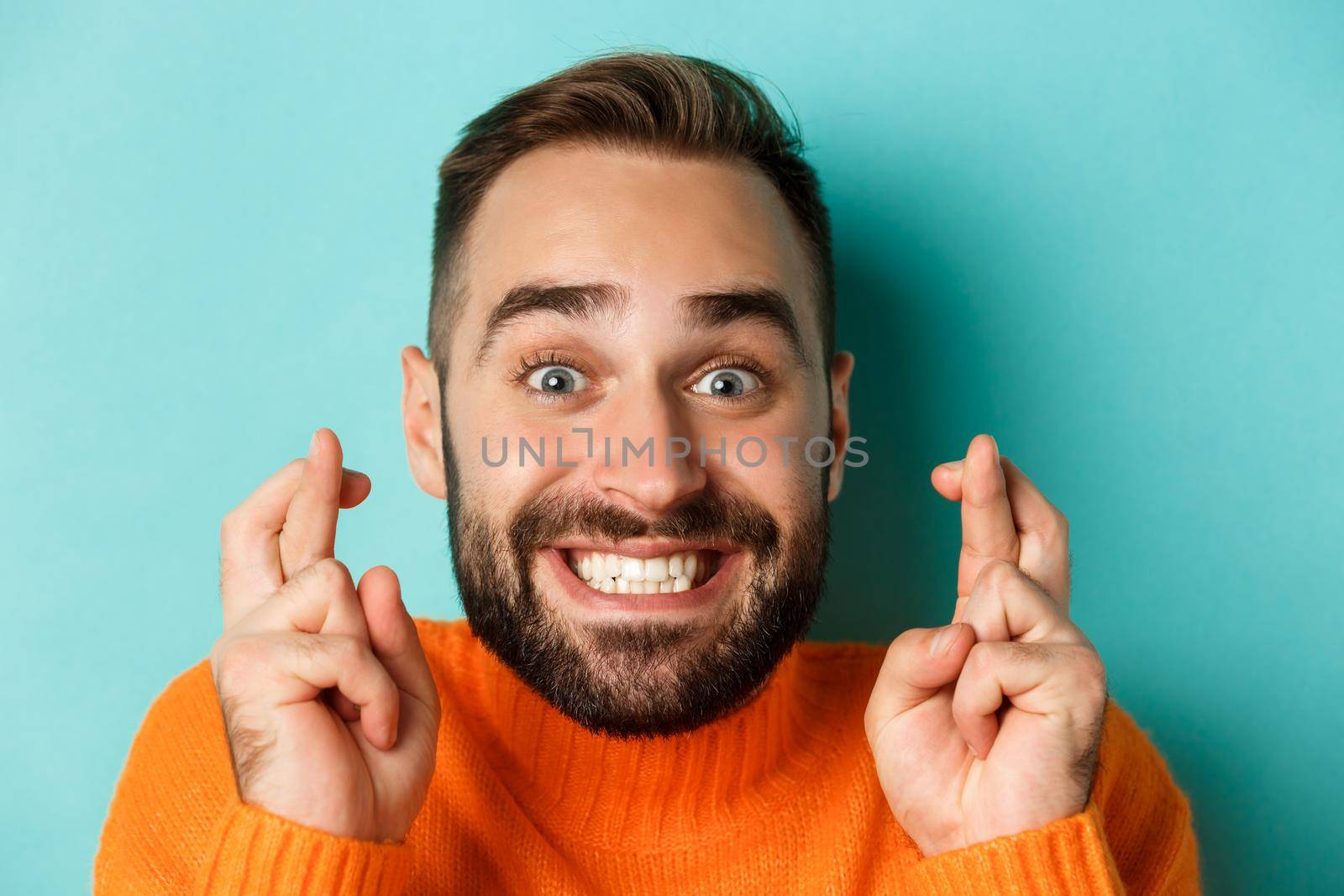 Headshot of hopeful bearded man making a wish, smiling and holding fingers crossed for good luck, standing over light blue background by Benzoix