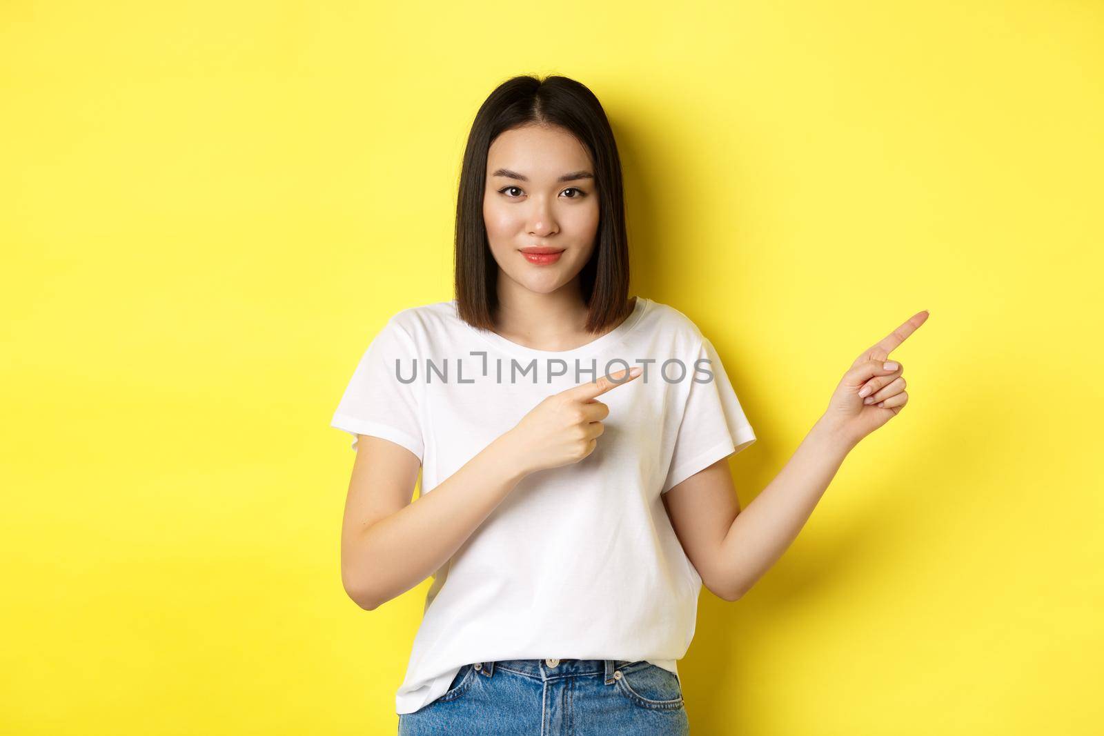 Beauty and fashion concept. Beautiful asian woman in white t-shirt pointing fingers right, demonstrate logo standing over yellow background by Benzoix