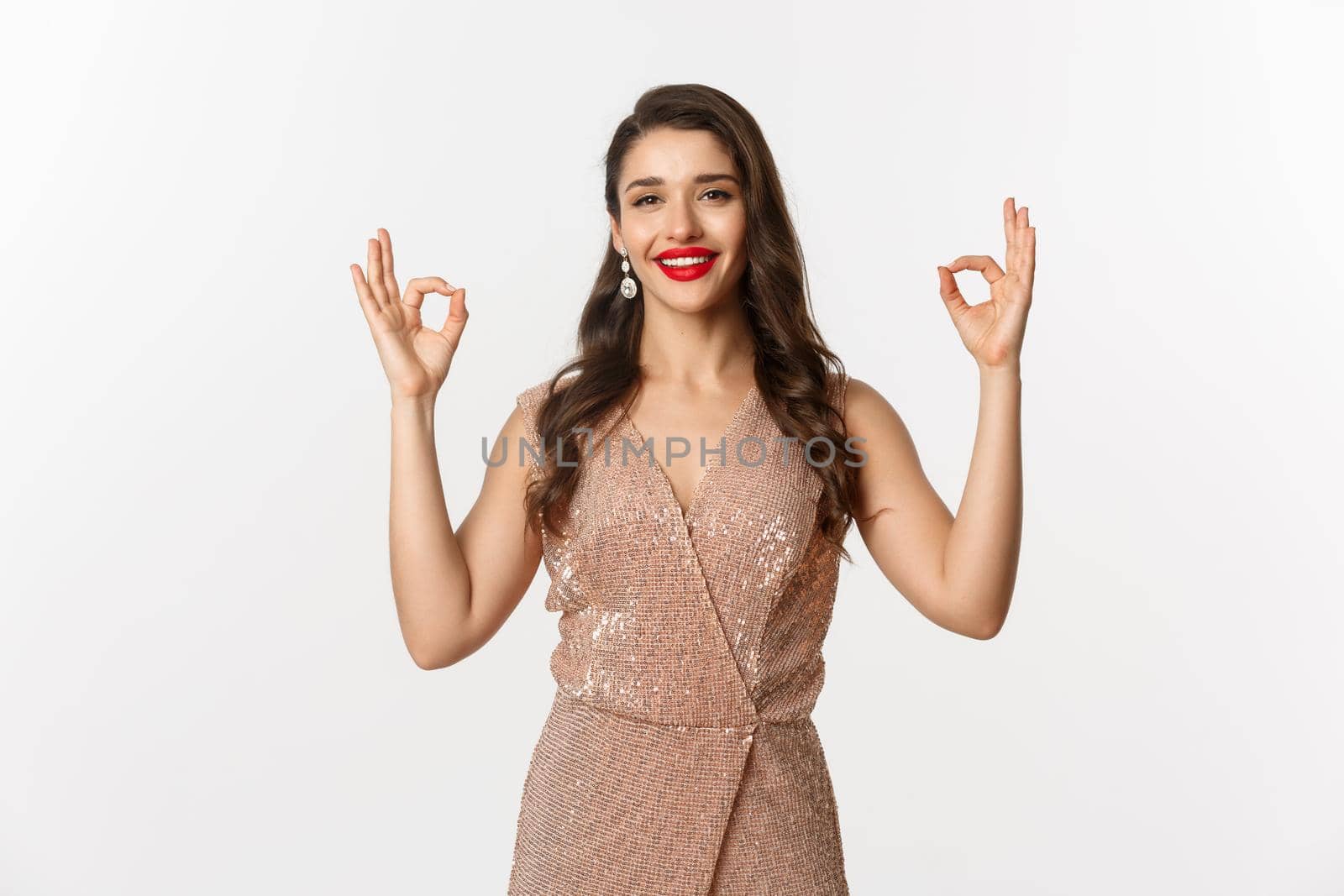 Portrait of satisfied beautiful woman recommending something, showing okay signs and smiling in approval, agree or like promo, standing in luxury party dress, white background.