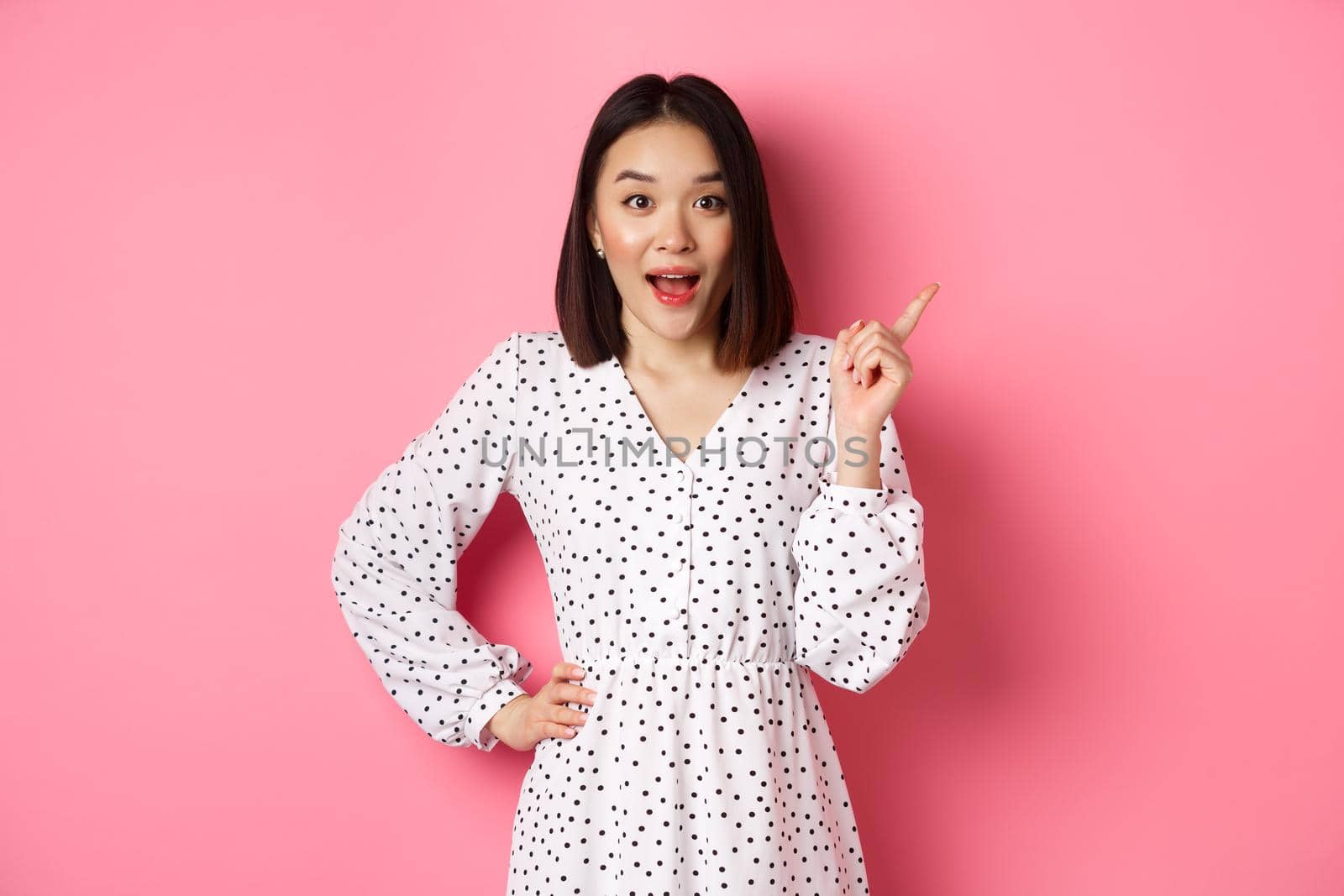 Excited pretty asian girl pointing at upper right corner, gasping amazed and looking at camera, discuss store promo offer, standing over pink background by Benzoix