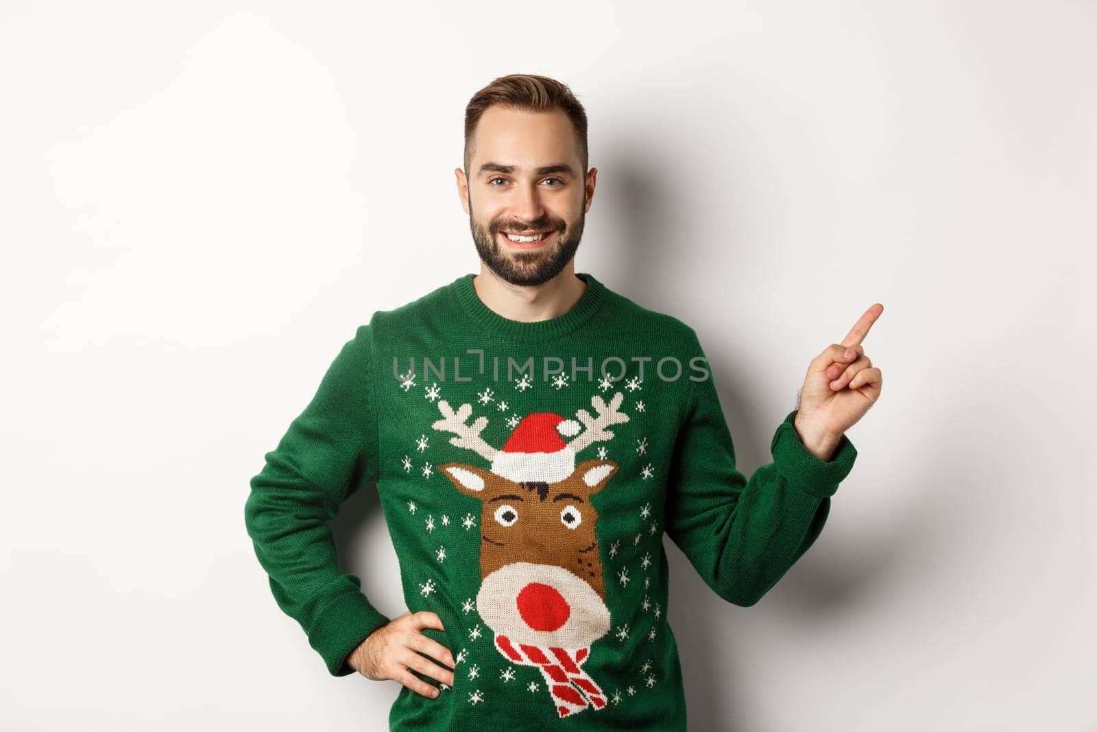 Winter holidays and christmas. Handsome bearded adult man pointing finger right at promo banner, standing in funny sweater against white background.