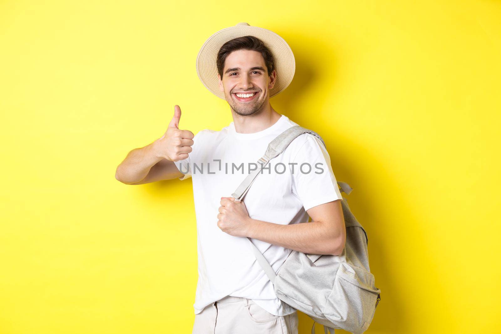 Tourism, travelling and holidays concept. Happy man going on vacation, holding backpack and showing thumb up, smiling satisfied, standing against yellow background.