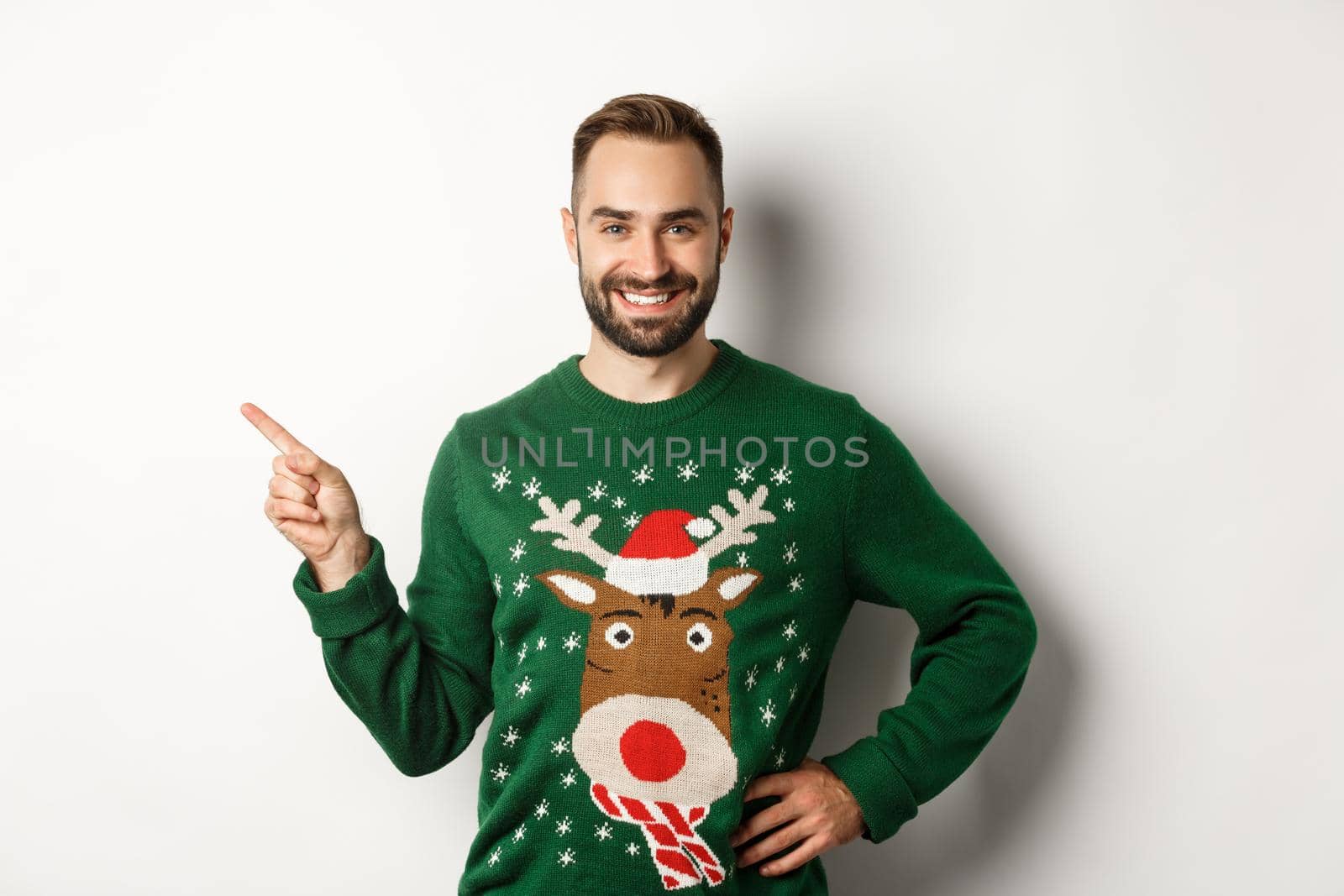 New year celebration and winter holidays concept. Confident and happy man with beard, wearing christmas sweater, pointing at upper left corner banner, white background.