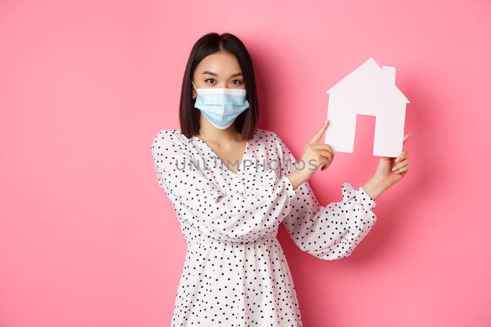 Covid-19, real estate and lifestyle concept. Cute asian woman in face mask selling houses, showing model of home and looking at camera, standing over pink background.