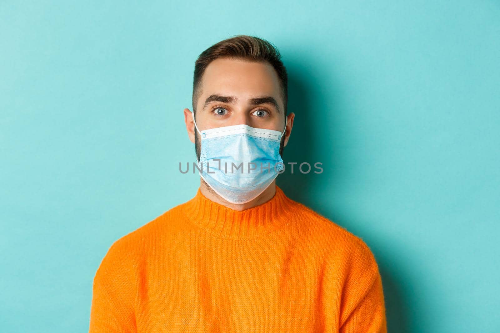 Covid-19, social distancing and quarantine concept. Close-up of young man in face mask staring at camera, standing over turquoise background.