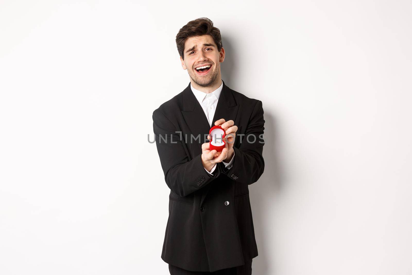 Portrait of handsome man in black suit, open box with wedding ring, making a proposal, asking to marry him, standing against white background.