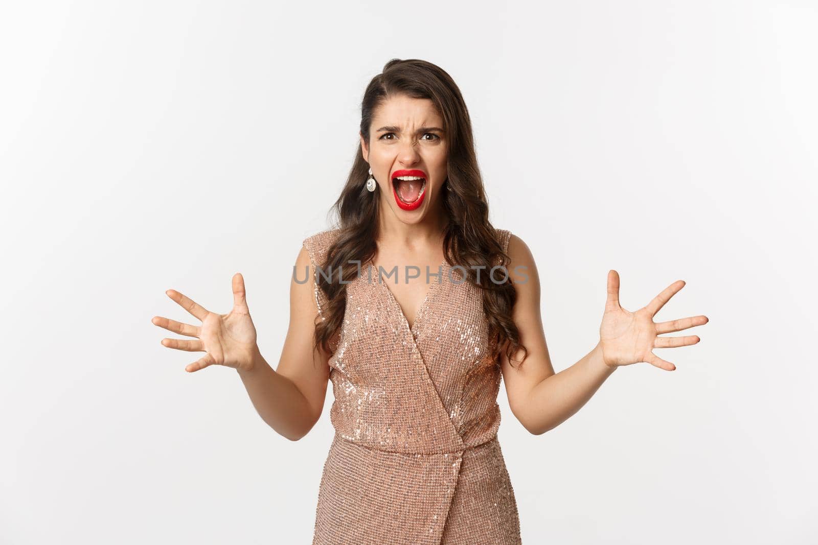 Celebration and party concept. Angry young woman in glamour dress shouting at you, looking mad and outraged, shaking hands aggressive, having an argument, white background.