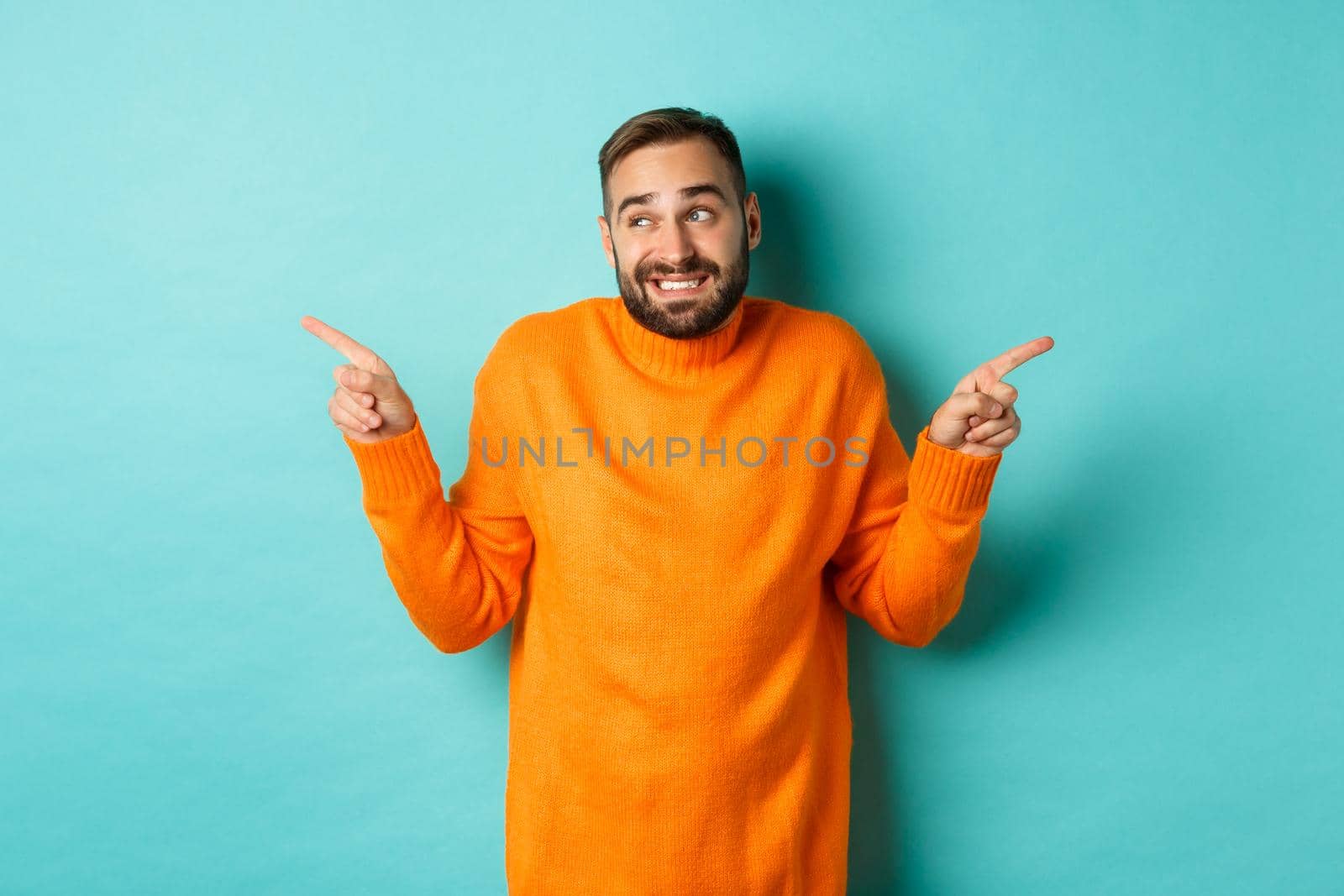 Confused handsome man shrugging indecisive, pointing fingers sideways, cant choose between two variants, standing against light blue background.