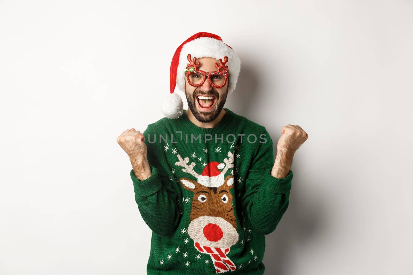 Christmas holidays, celebration concept. Happy man in Santa hat triumphing, wearing funny party glasses and rejoicing, standing over white background.