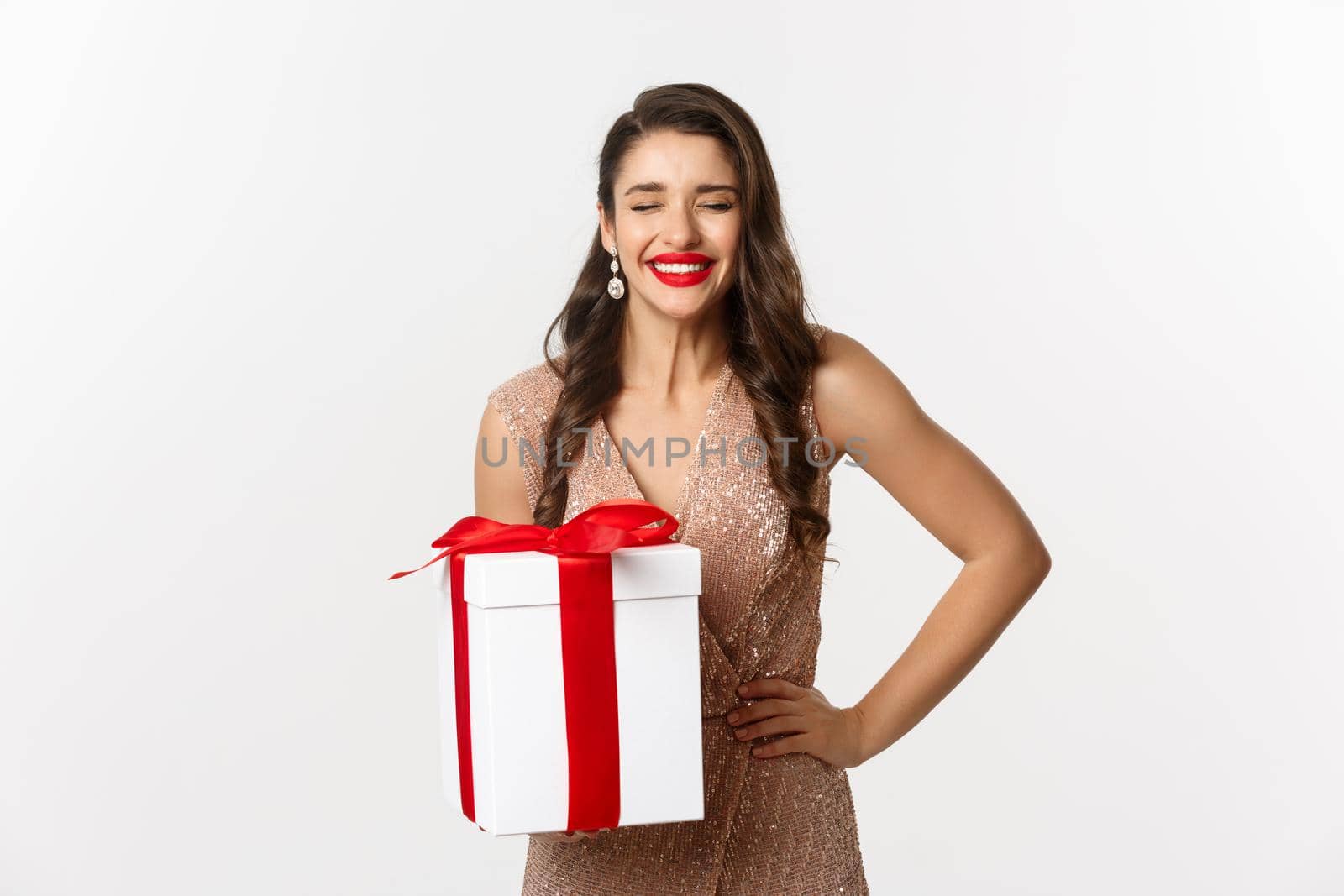 Merry Christmas. Image of attractive woman in luxury dress, celebrating winter holidays, holding gift and laughing from happiness, standing over white background by Benzoix