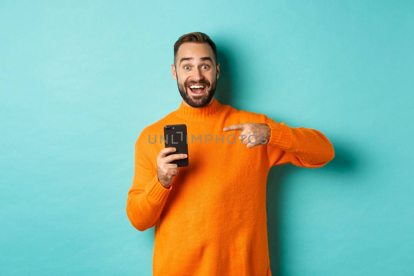 Excited man pointing at mobile phone, showing internet promo, standing over turquoise background, concept of online shopping by Benzoix