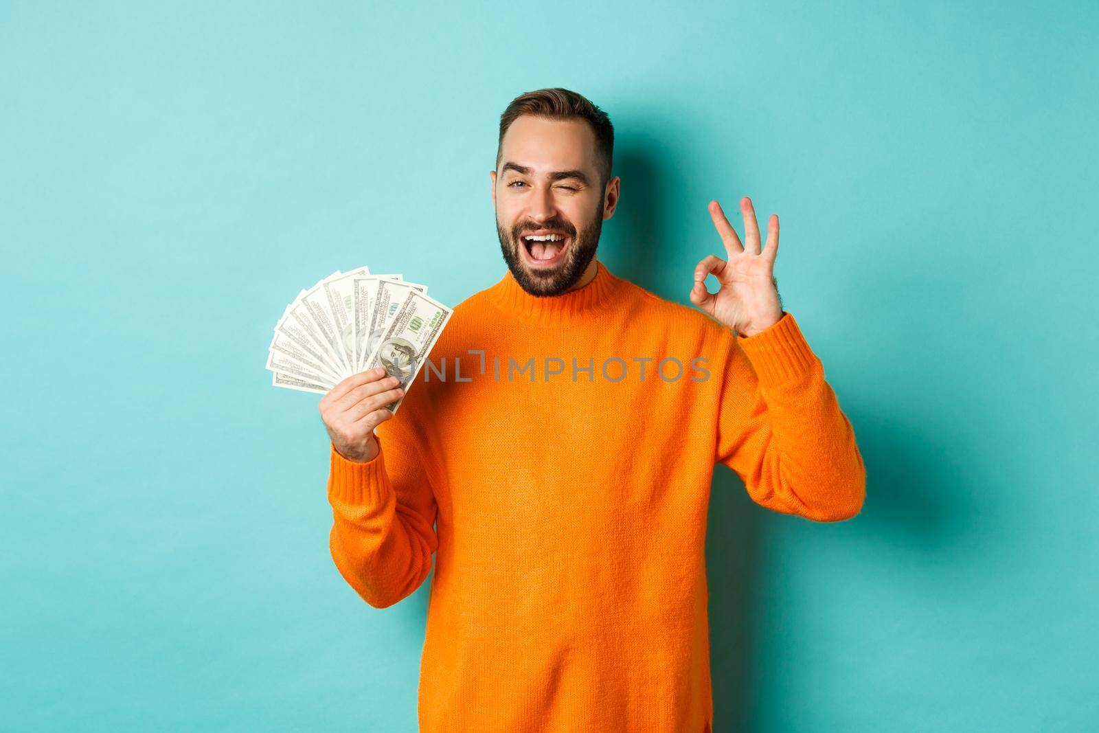 Shopping. Excited guy holding money, showing ok sign and winking, standing over light blue background. Copy space