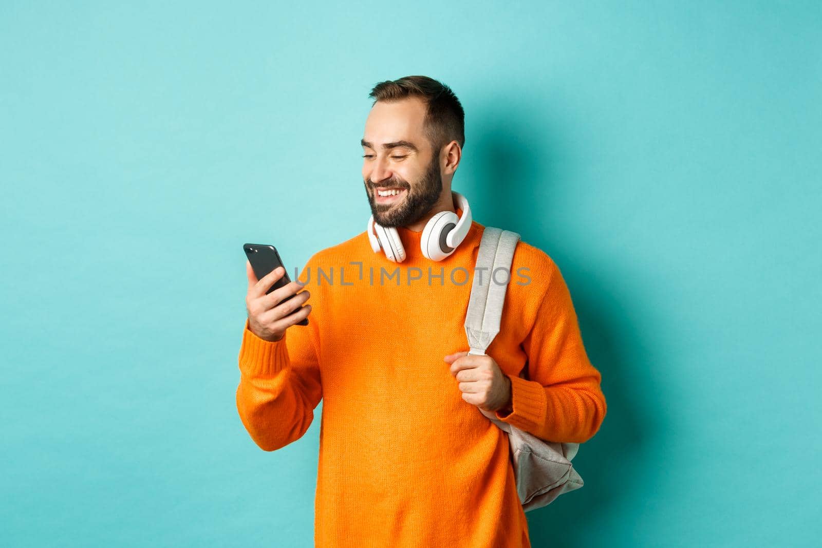 Caucasian man with headphones and backpack looking at phone, reading message and smiling, standing over turquoise background by Benzoix