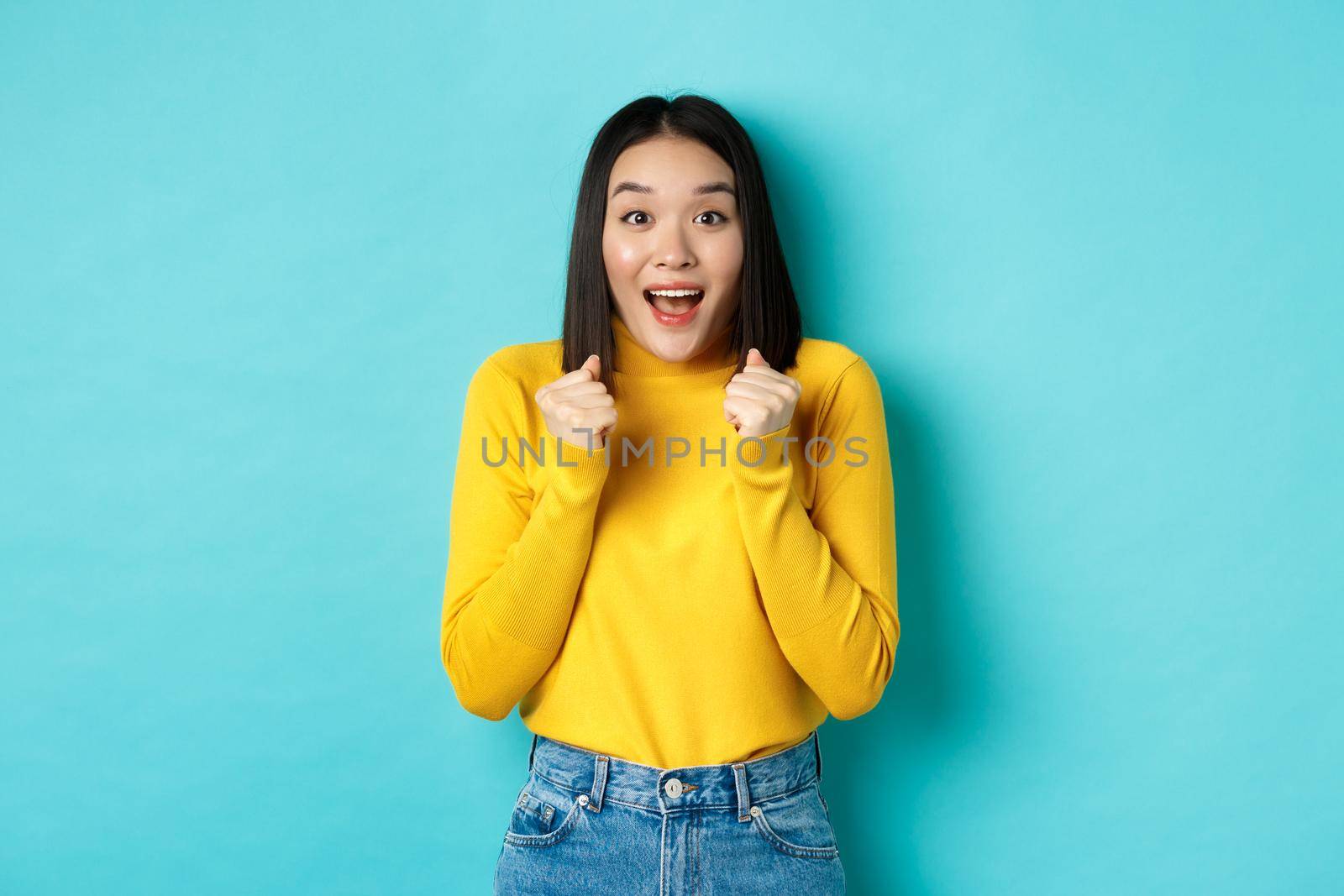 Hopeful and happy asian woman looking with amazement and hope at camera, winning, standing over blue background.