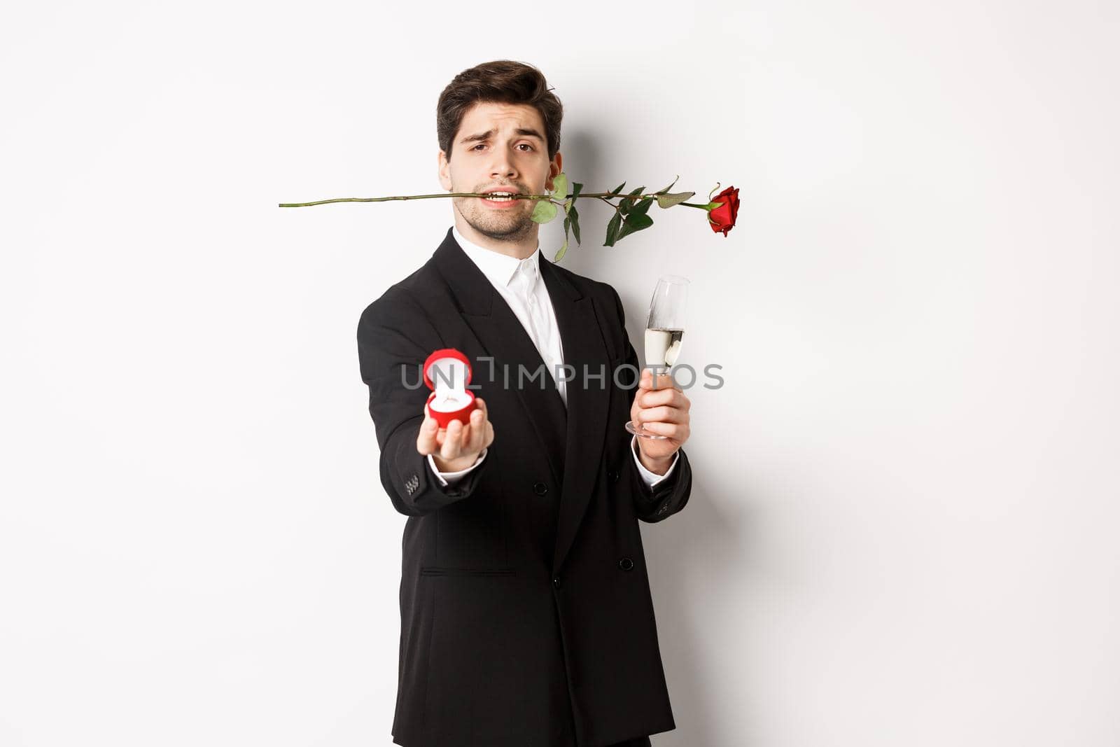 Romantic young man in suit making a proposal, holding rose in teeth and glass of champagne, showing engagement ring, asking to marry him, standing against white background by Benzoix