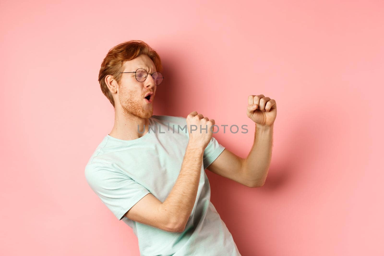 Cheerful redhead guy listening music and dancing, triumphing or celebrating success, standing over pink background.