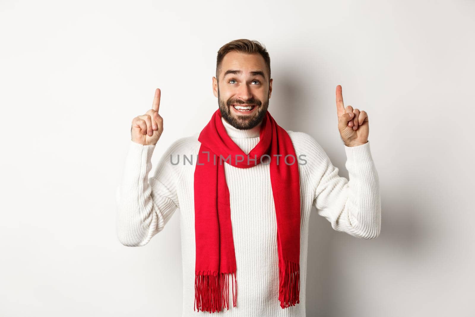 Christmas shopping and winter holidays concept. Happy bearded man in red scarf pointing fingers up, looking excited at copy space, standing over white background.