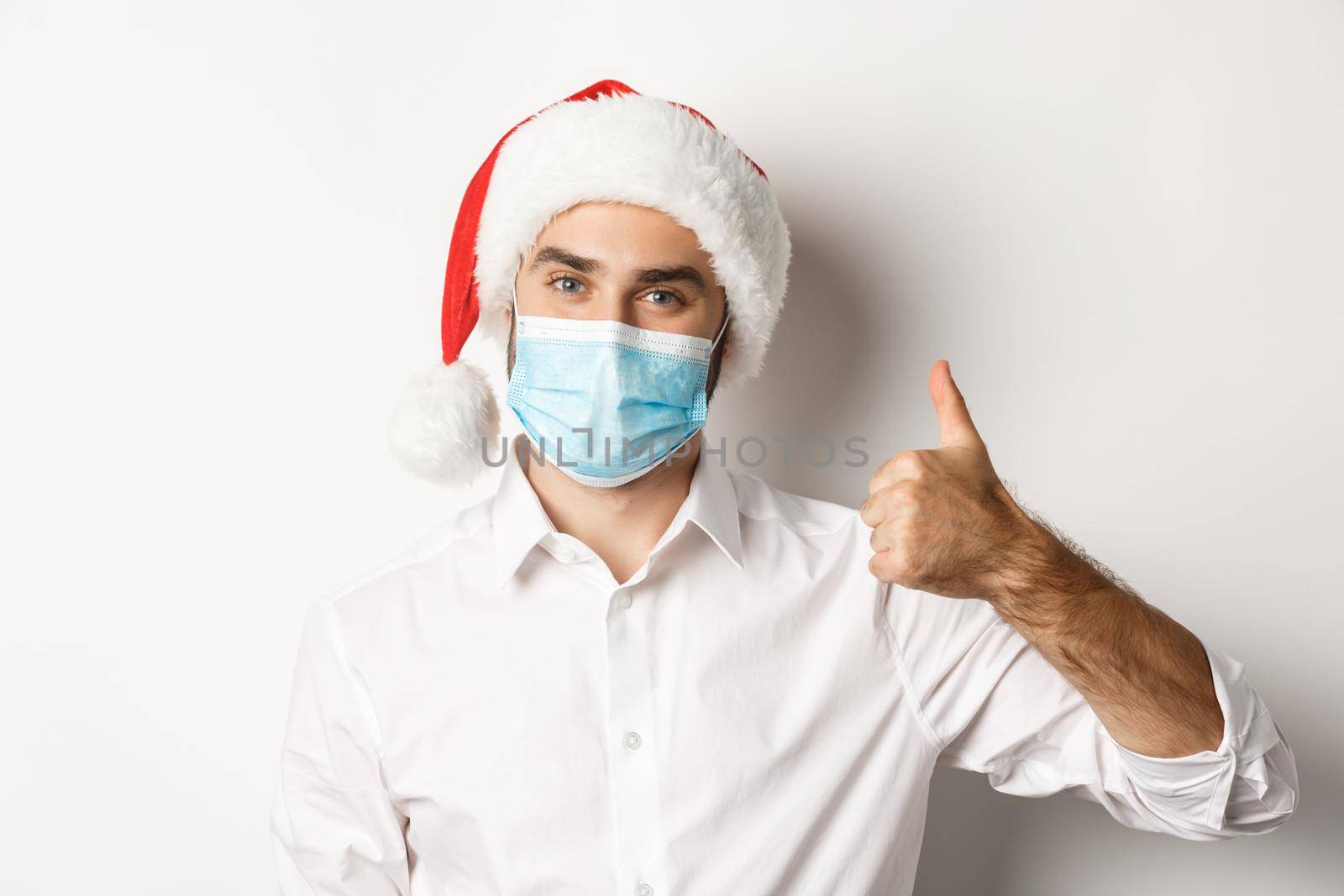 Concept of covid-19, social distancing and winter holidays. Satisfied man in face mask and santa hat showing thumb up, celebrating christmas with preventive measures, white background.
