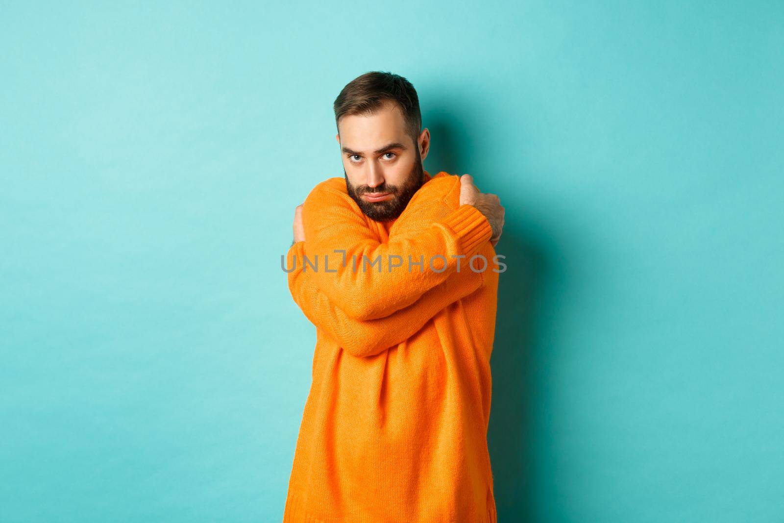 Timid man feeling offended and defensive, hugging himself and looking suspicious at camera, standing over light blue background by Benzoix