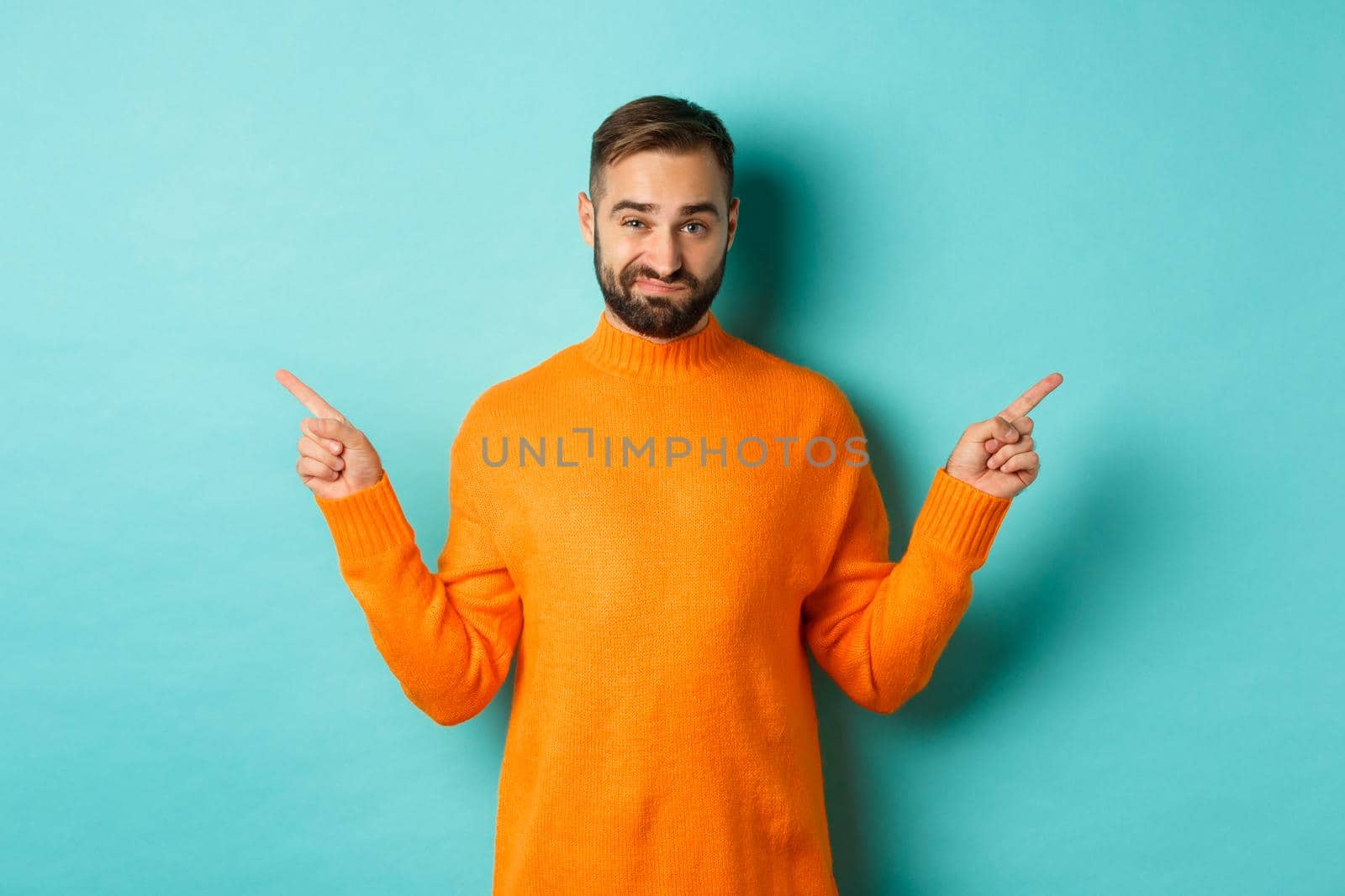 Doubtful and indecisive man pointing fingers sideways, showing different sides, two choices, looking skeptical, standing over light blue background.