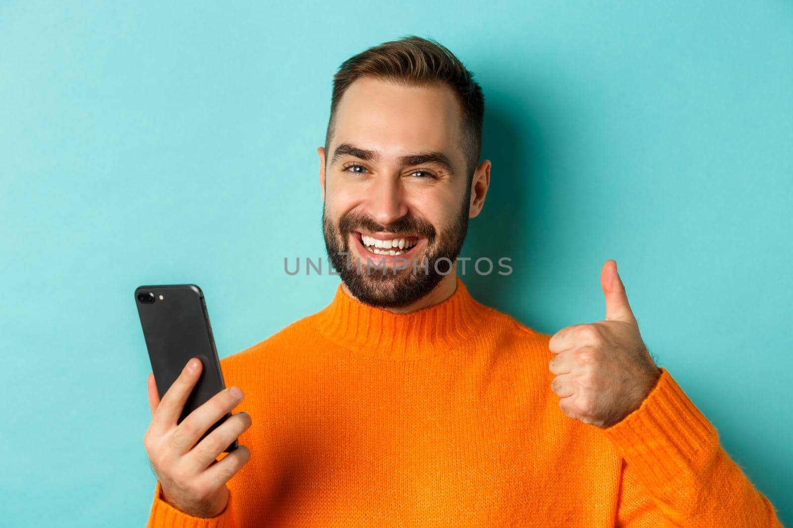 Close-up of bearded man holding smartphone, thumbs up, recommending mobile app, standing satisfied over turquoise background.