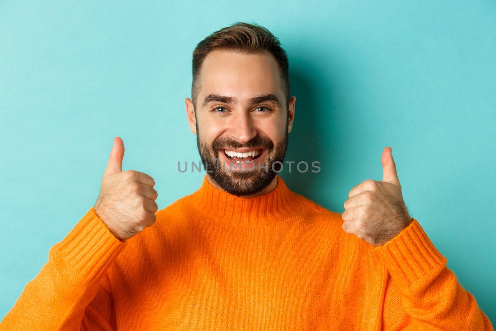 Close-up of attractive bearded man showing thumb-up, approve and like, recommending product, light blue background.