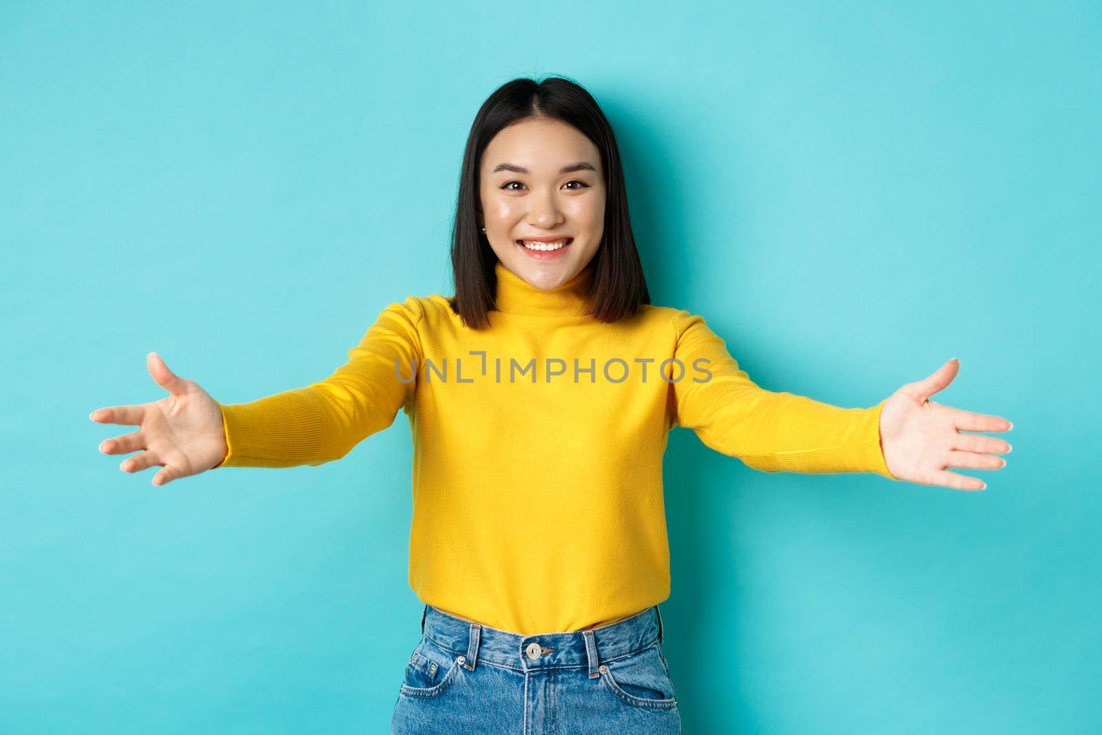 Portrait of beautiful korean woman spread out hands for hug, reaching for cuddles and smiling at camera, greeting you, standing over blue background by Benzoix