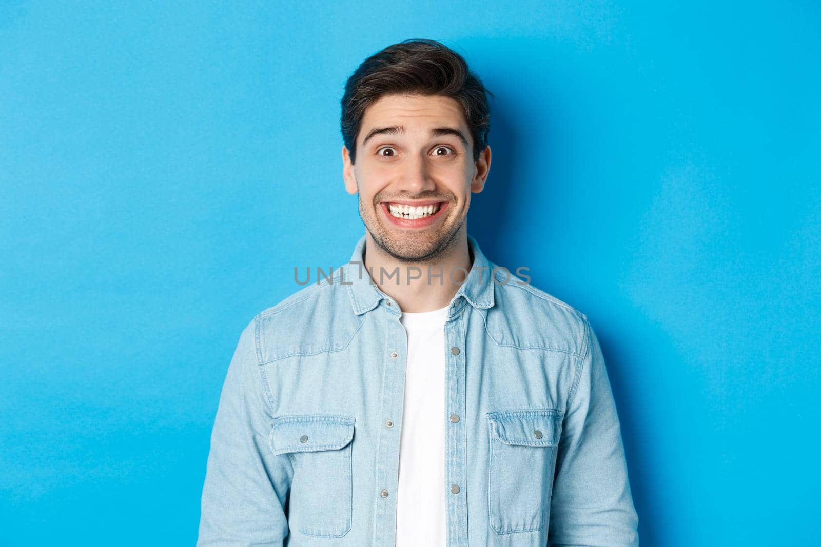 Close-up of smiling excited man with beard, looking amused at advertisement, standing against blue background by Benzoix