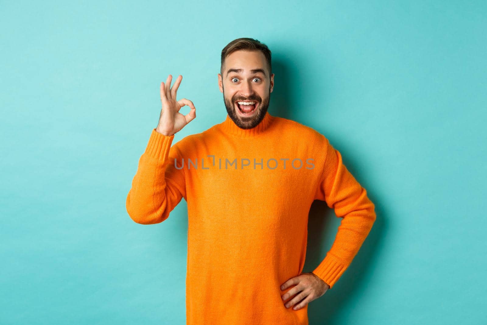 Impressed man recommending promo, showing okay sign and smiling amazed, approve and agree, standing over light blue background.