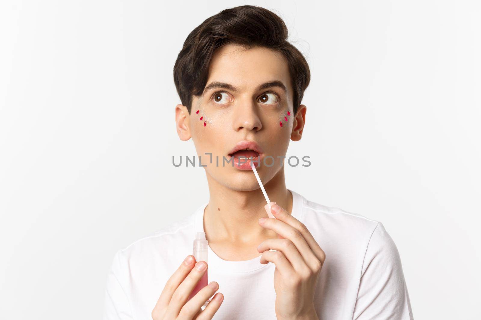 Close-up of beautiful androgynous man with glitter under eyes applying lip gloss, looking dreamy at upper left corner, white background by Benzoix