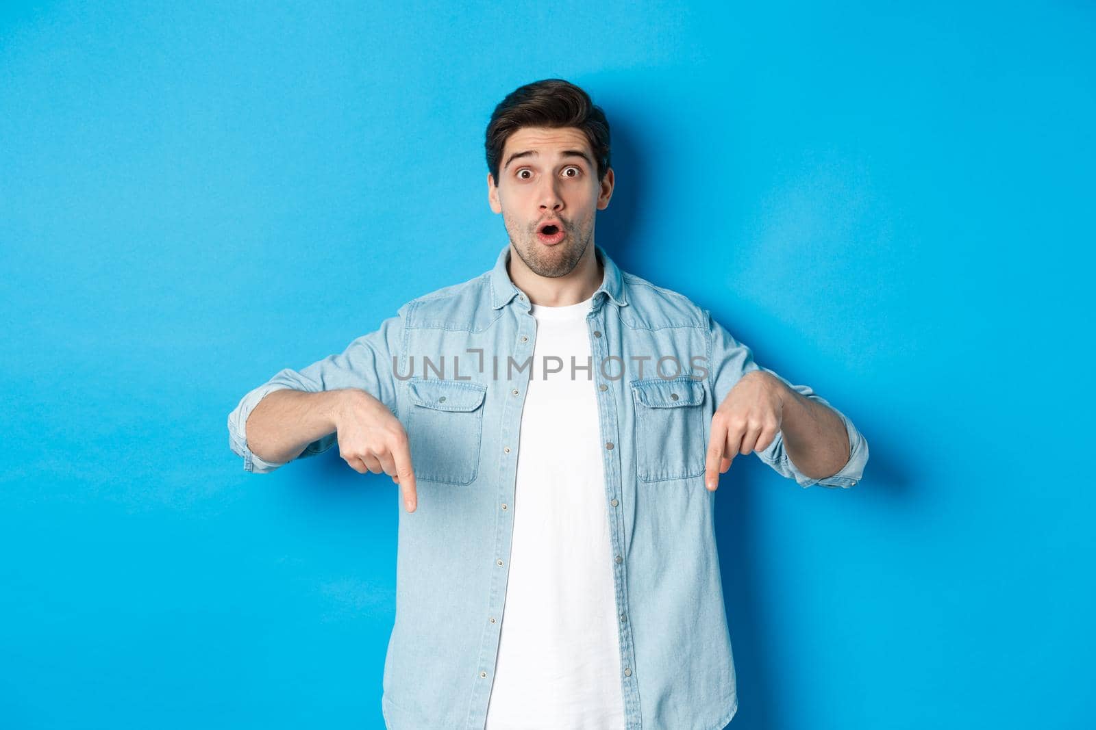 Surprised bearded adult man in casual outfit, pointing fingers down and looking startled, asking question about promotion, standing over blue background by Benzoix