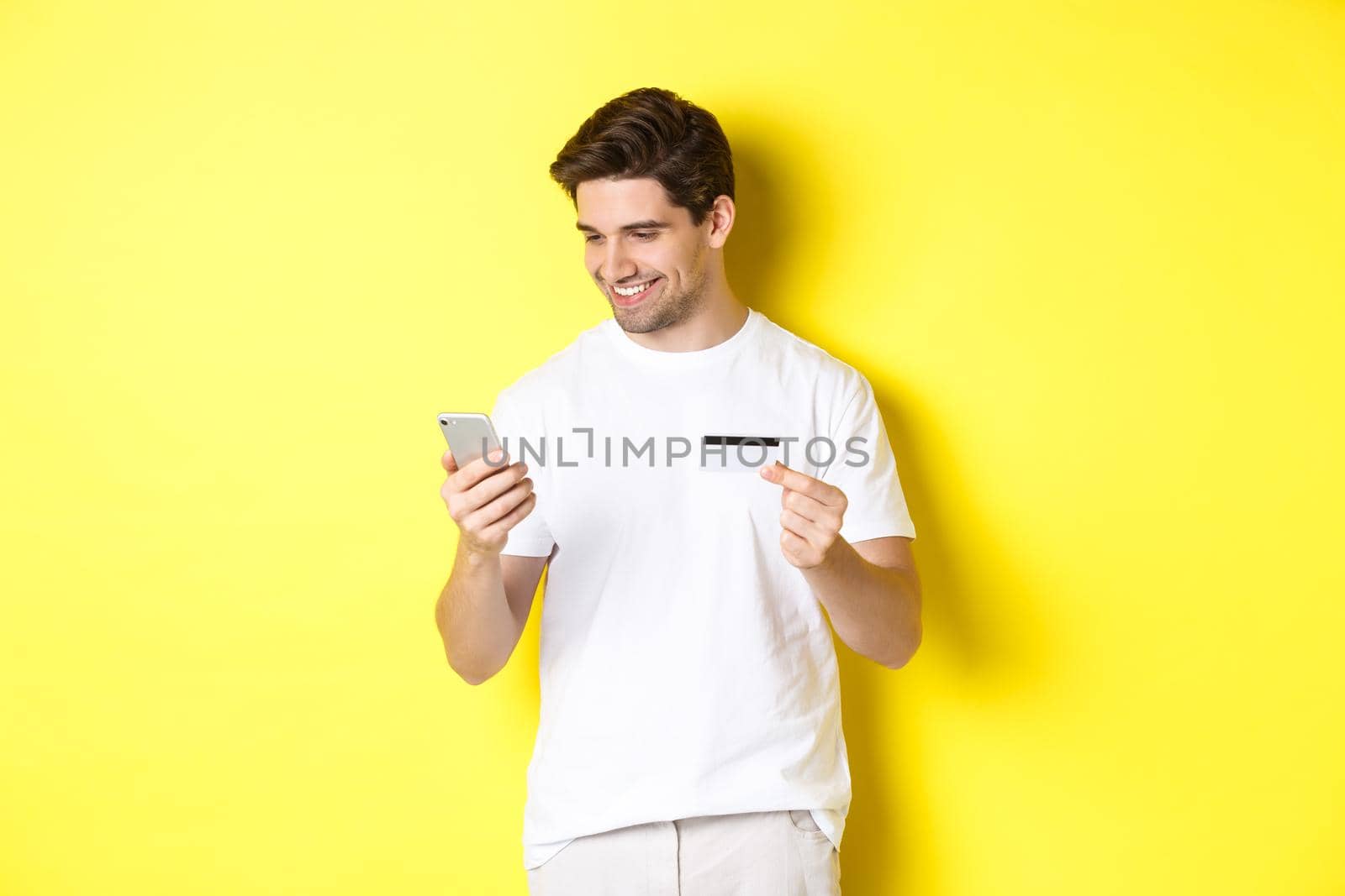 Young man paying online, insert credit card number on mobile phone, shopping in internet, standing over yellow background.