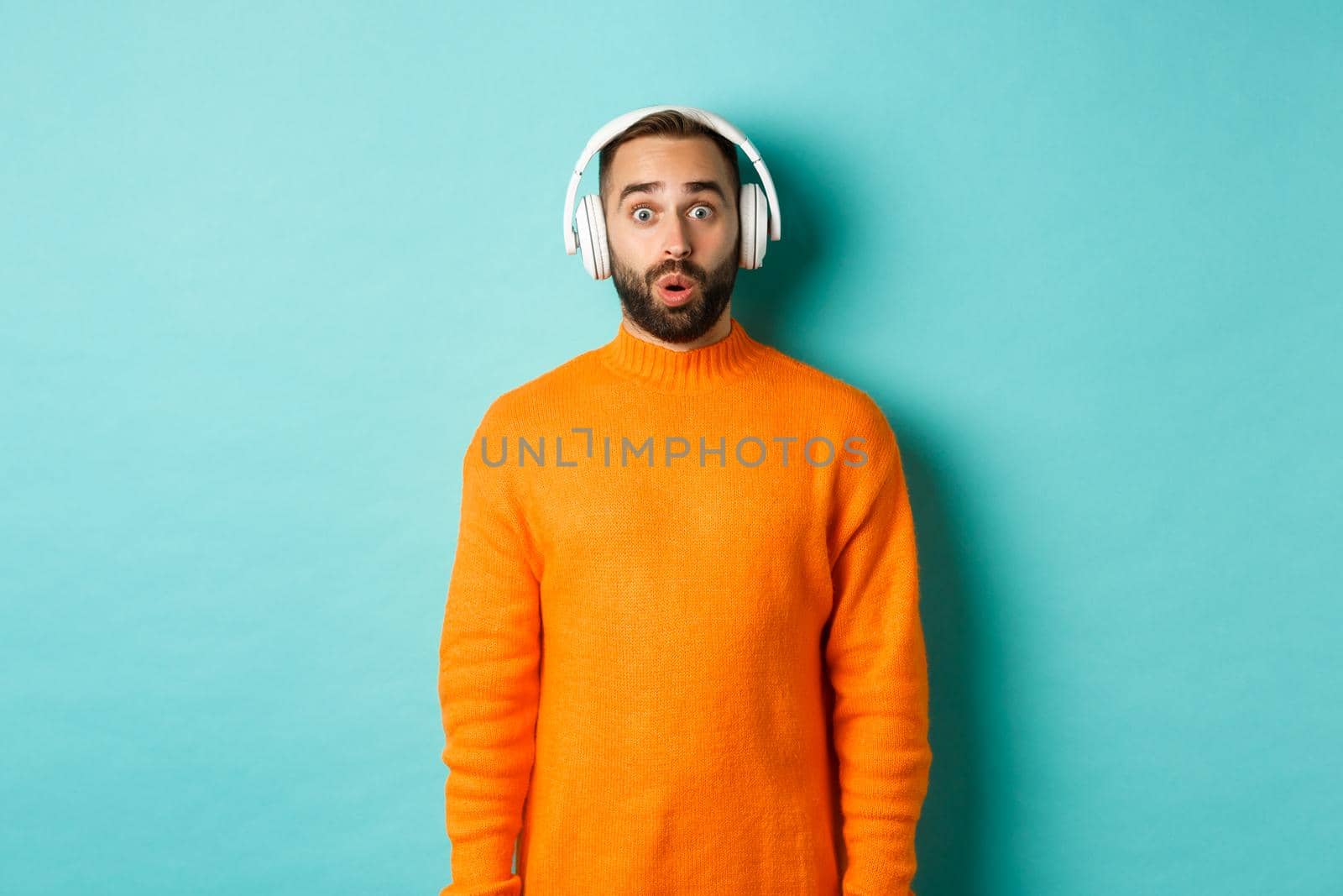 Amazed adult man listening music in headphones, looking at camera impressed with sound, standing over turquoise background.